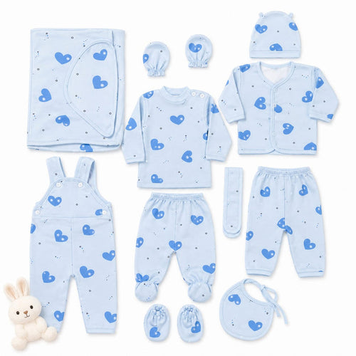 A baby clothing set laid out on a white background, including a light blue long-sleeve top, matching footed pants, overalls, a cardigan, a bonnet, a bib, a hat, a muslin cloth, and booties, all printed with a blue heart pattern. A small plush bunny toy is