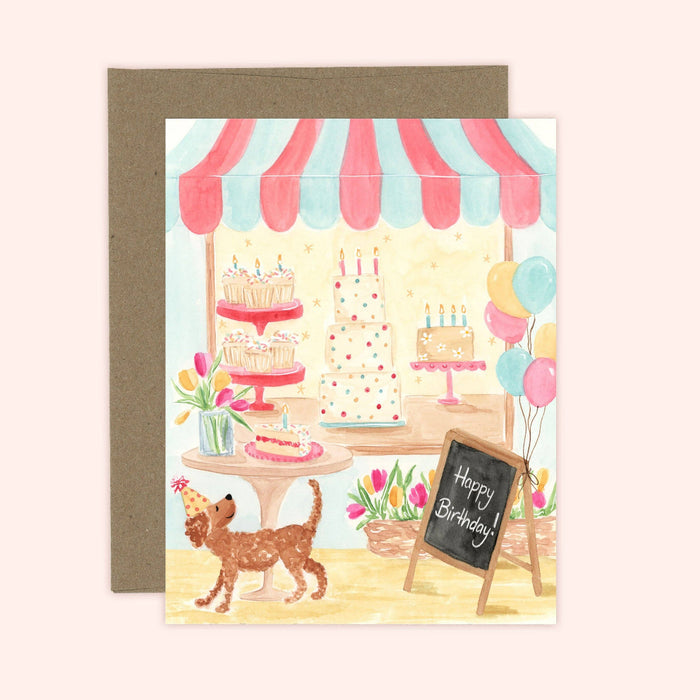 Birthday card with a dog, cake stand, and balloons in front of a cake shop.