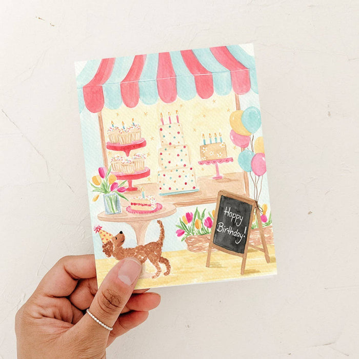 Hand holding a birthday card with a cake stand and dog illustration on a white background