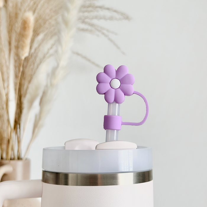 A close-up of a white tumbler with a silver rim, showcasing the Coconut Lane Lilac Flower Straw Cover. In the background, ornamental pampas grass stems are placed in a vase, slightly blurred to enhance the light and airy ambiance.