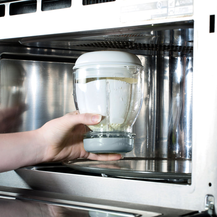 A hand places a glass container with a white lid from the Mighty Blender 22-Piece Baby Food System into a stainless steel microwave, preparing homemade baby food. The container appears to hold water.