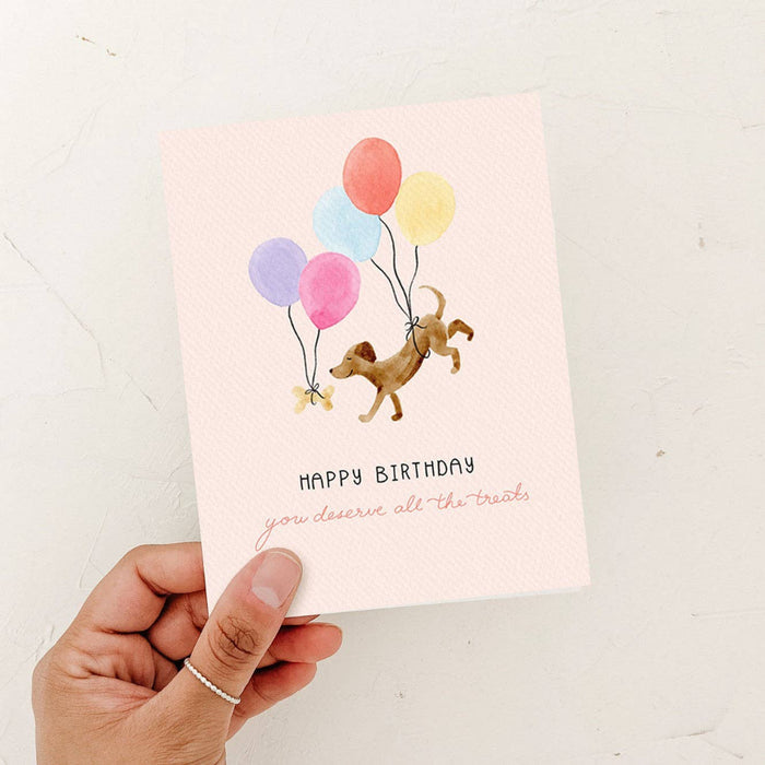 Hand holding a birthday card with a dog and balloons illustration on a light background